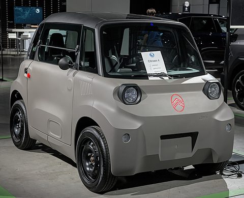 Taupe cube-shaped two-seater with new round headlamps, a red chevron badge and black steel wheels on a show floor.