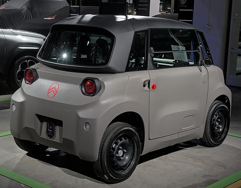 Rear of a taupe cube-shaped two-seater with round red tail lamps and a red chevron badge, on a show floor. Opens a larger view
