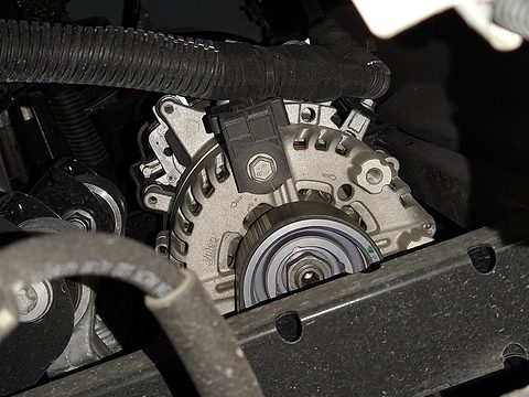 Close view of a finned silver electric motor with a belt pulley, among cables under a small car. Opens a larger view