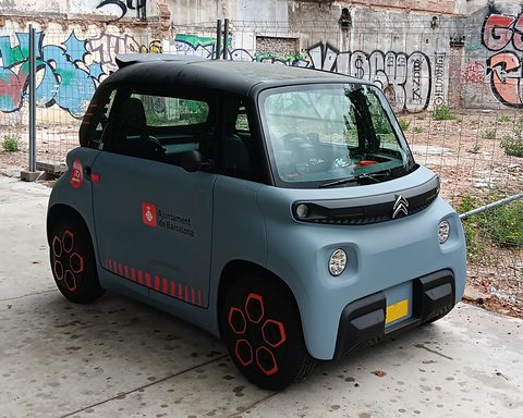 Grey cube-shaped two-seater with orange wheels and Ajuntament de Barcelona lettering, parked by a fence before a graffiti wall. Opens a larger view