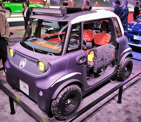 Lilac and white open two-seater with yellow lamps, a roof light bar and chunky tyres, raised on a show stand. Opens a larger view