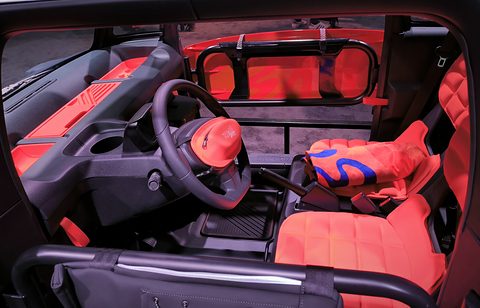 Open cabin with quilted orange seats, a small steering wheel and black dashboard, seen from the side. Opens a larger view