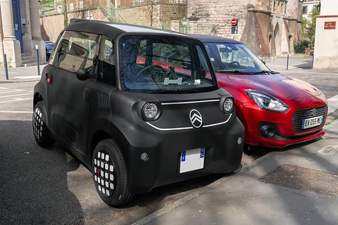 Black cube-shaped two-seater with white-dotted wheel covers parked beside a red hatchback on a sunny street. Opens a larger view