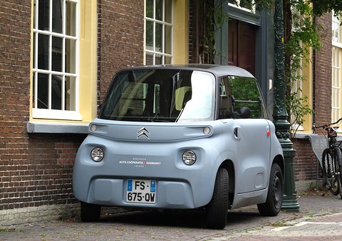 Grey cube-shaped two-seater with round lamps and a French plate, parked on a pavement in front of a brick façade with tall windows. Opens a larger view