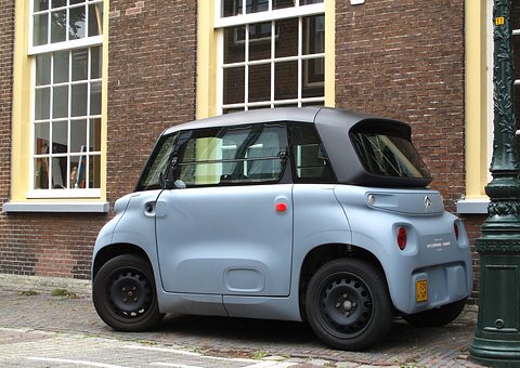 Rear of a grey cube-shaped two-seater with black wheels, parked by a brick house and a green lamp post. Opens a larger view