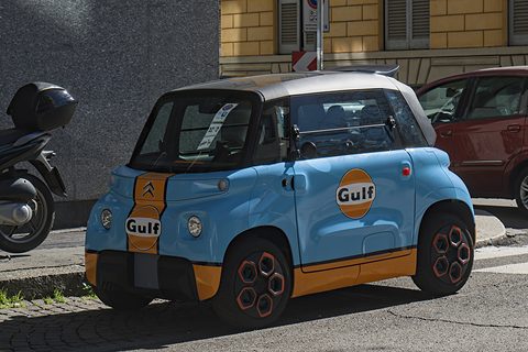 Pale blue cube-shaped two-seater with an orange stripe and Gulf roundels, parked at a kerb beside a scooter. Opens a larger view