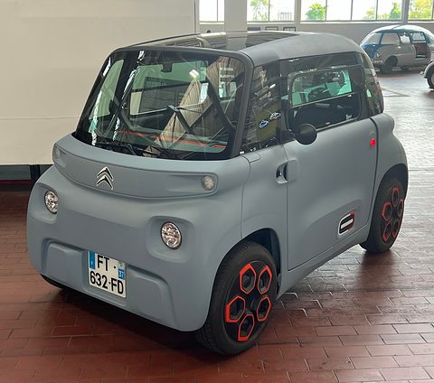 Grey cube-shaped two-seater with red wheel inserts and a French plate on a brick museum floor. Opens a larger view
