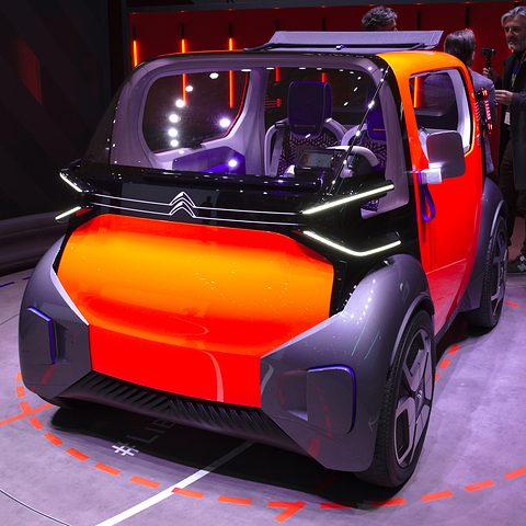 Front of an orange and dark grey box-shaped concept car with a thin light bar and a glazed cabin, on a dark stand. Opens a larger view