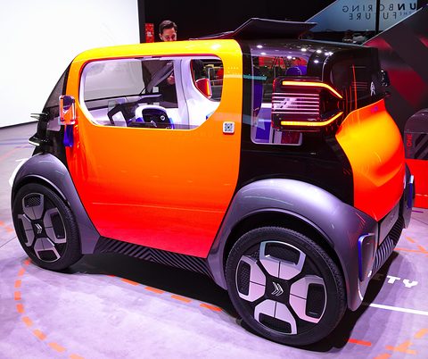 Orange and dark grey cube-shaped two-seater with large wheels and a lit tail lamp, on a stand marked with orange lines.