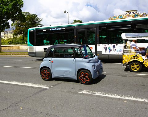 Grey cube-shaped two-seater with orange wheel inserts on a wide road, a green and white city bus and a tourist trike behind.