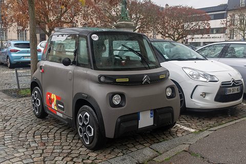 Taupe cube-shaped two-seater with an orange and red number graphic on its door, parked on cobbles beside a white hatchback. Opens a larger view