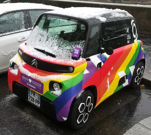 Small cube-shaped car in diagonal rainbow stripes with snow on its roof and bonnet, parked beside a white car. Opens a larger view