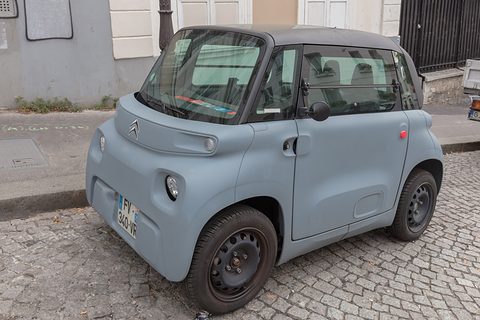 Grey cube-shaped two-seater with black steel wheels parked on granite setts by a pavement. Opens a larger view