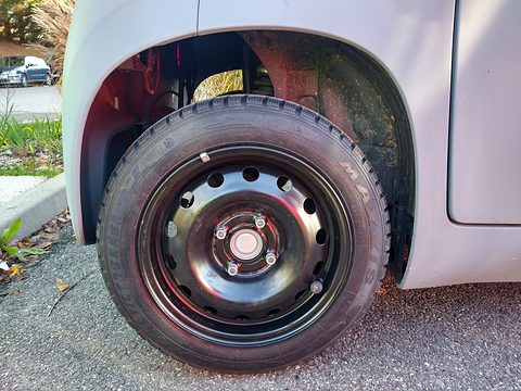 Black steel wheel and winter tyre under the grey wheel arch of a small car parked on gravel. Opens a larger view