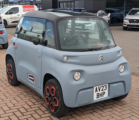 Grey cube-shaped two-seater with orange wheel inserts and a British plate, parked on brick paving by a dealer’s vans. Opens a larger view