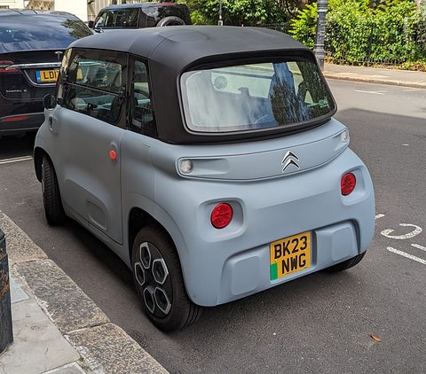 Rear of a grey cube-shaped two-seater with round red tail lamps and a British plate, parked beside a pavement. Opens a larger view