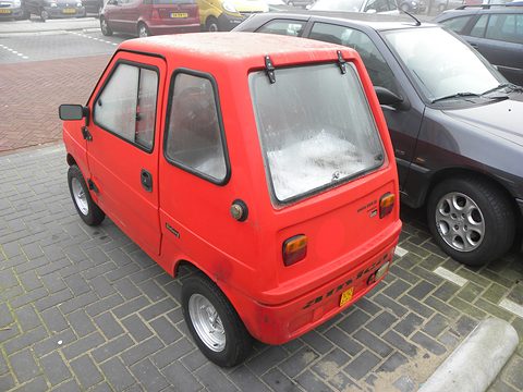 Red boxy microcar seen from the rear left, with a large hatch window and a yellow Dutch plate, beside a grey hatchback. Opens a larger view