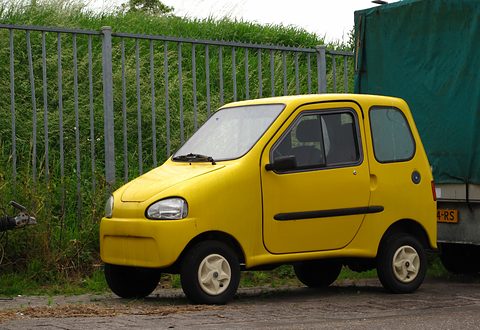 Small rounded yellow two-door microcar with cream wheels, parked by a steel fence in front of tall grass. Opens a larger view