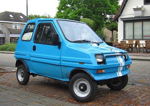 Blue boxy two-door microcar with twin white stripes over its nose and a chrome grille bar, parked on a brick street. Opens a larger view