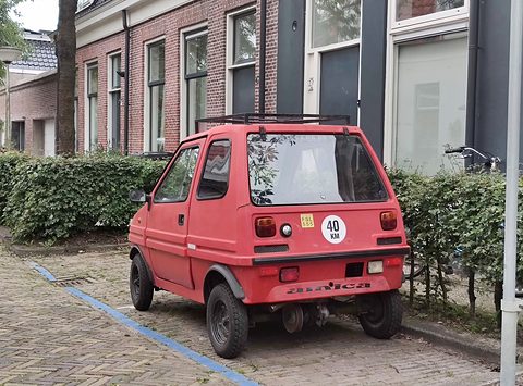 Rear of a red boxy microcar with amica lettering on the bumper, a roof rack and a round speed disc, parked on brick paving. Opens a larger view