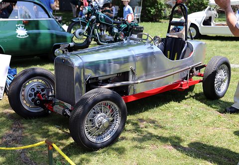 A narrow polished single-seat racing car with a red chassis and wire wheels on a lawn, motorcycles and cars behind. Opens a larger view