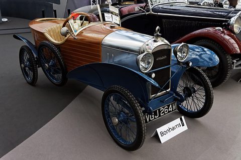 A small car with a planked wooden body, a polished bonnet, blue wings and wire wheels, a sale sign before it. Opens a larger view