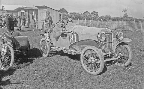 A goggled driver in a mud-spattered cyclecar holding a bouquet, a motorcycle sidecar numbered 44 beside it on grass, sheds behind.
