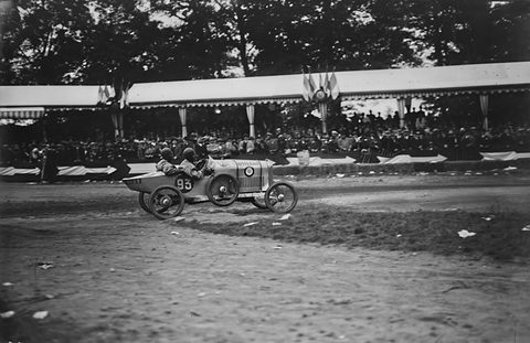 A small racing cyclecar numbered 93 speeds along a dirt track in front of a long covered grandstand full of spectators. Opens a larger view