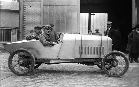 Side view of a long, low aluminium-bodied racing cyclecar with a pointed tail, two men aboard, on cobbles before a workshop. Opens a larger view
