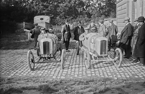 Two narrow racing cyclecars facing the camera on cobbles, drivers in caps, a group of men in overcoats behind. Opens a larger view