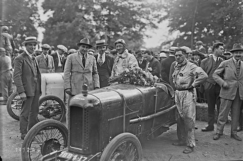 A dark racing two-seater with a flat radiator and wire wheels, flowers on its scuttle, men in suits and overalls standing round it. Opens a larger view