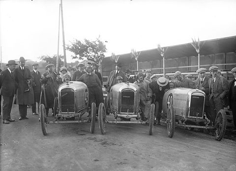 Three small racing cyclecars with pointed radiators lined up nose-first before a crowd of men in caps and coats. Opens a larger view