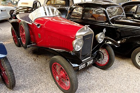 A red open roadster with black wings and red wire wheels on a gravel floor beside a black saloon. Opens a larger view