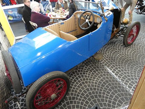 Looking down into a blue open car with tan seats, a black steering wheel and red wire wheels, people at a table behind. Opens a larger view