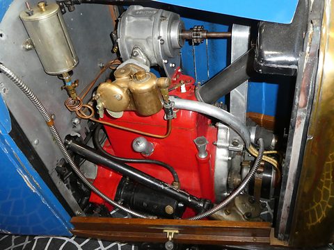 A red four-cylinder engine with a brass carburettor, cables and a polished oil tank in a blue bonnet bay. Opens a larger view