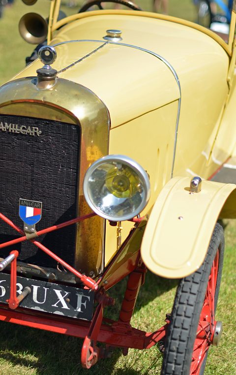 Close view of a brass radiator with the Amilcar name, a French flag badge, a chrome headlamp and a cream wing over a red wheel. Opens a larger view
