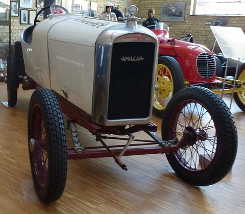 Front view of a white cyclecar with a black honeycomb radiator, maroon wire wheels and a red racing car behind. Opens a larger view