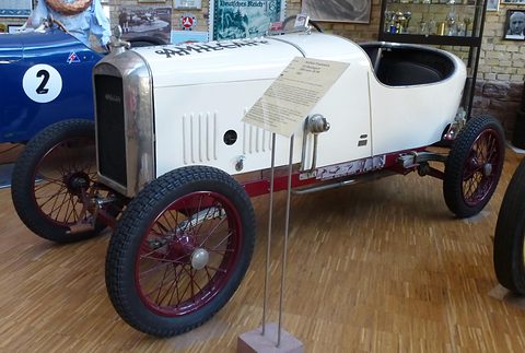 Side view of a white cyclecar with bonnet louvres, maroon wire wheels and a pointed tail, a label on a stand in front of it.