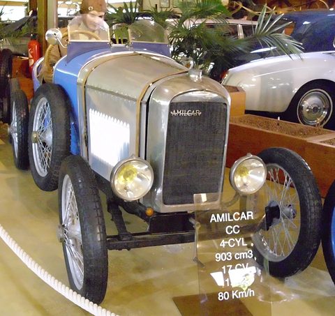 A small car with polished bonnet and blue wings, a clear placard listing its engine before it, palms and other cars behind. Opens a larger view