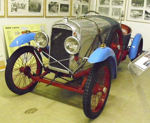 A small open car with a polished radiator, large headlamps, light blue wings and red wire wheels, framed photographs on the wall behind. Opens a larger view