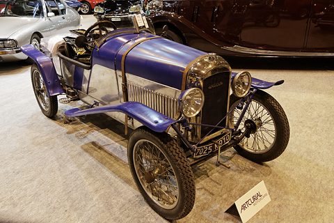 A narrow two-seat sports cyclecar with a polished aluminium body, purple-blue wings and wire wheels on a show carpet. Opens a larger view