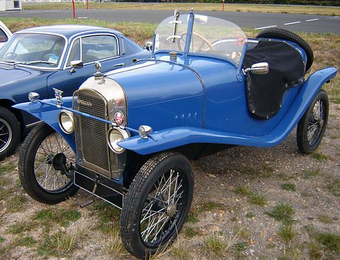 A small blue two-seater with a mesh radiator, cycle wings, wire wheels and a black tonneau over the cockpit. Opens a larger view