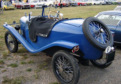 Rear three-quarter view of a small blue two-seater with a spare wire wheel on its pointed tail and a black tonneau. Opens a larger view