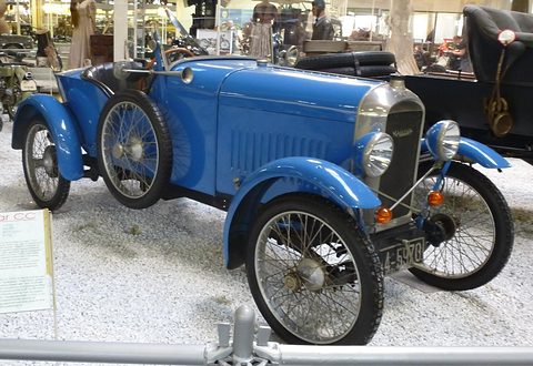 A pale blue cyclecar with a chrome radiator, two headlamps and wire wheels, a barrier rail across the foreground. Opens a larger view