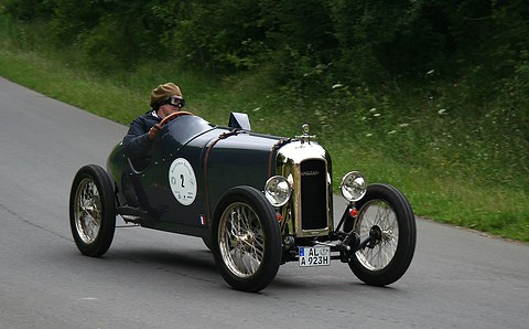 A dark green sports cyclecar with a polished radiator driven up a tarmac road by a goggled driver in a leather cap. Opens a larger view