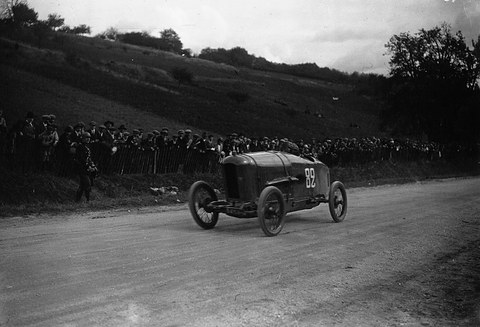 A small pointed-tail racing car with the number 82 climbing a dirt road, spectators lining a fence below a grassy hillside. Opens a larger view