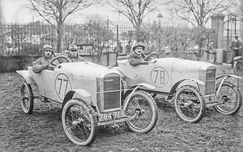 Two small pale cyclecars numbered 77 and 78 side by side on muddy grass, goggled drivers aboard, railings and trees behind. Opens a larger view