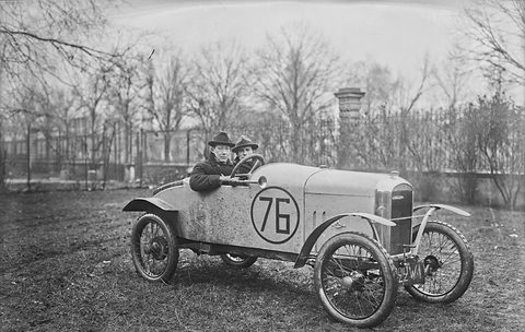 Two men in hats sit in a small light-coloured cyclecar with a big number 76 on its side, parked on rough grass before bare trees. Opens a larger view