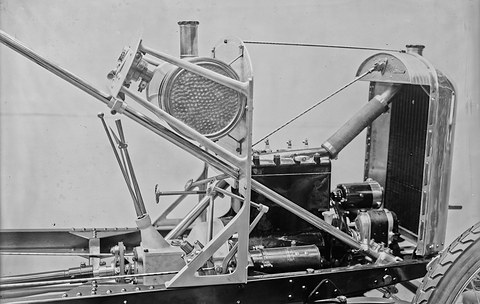 Black-and-white close view of a bare car chassis: a small four-cylinder engine, radiator, steering column and dashboard frame.