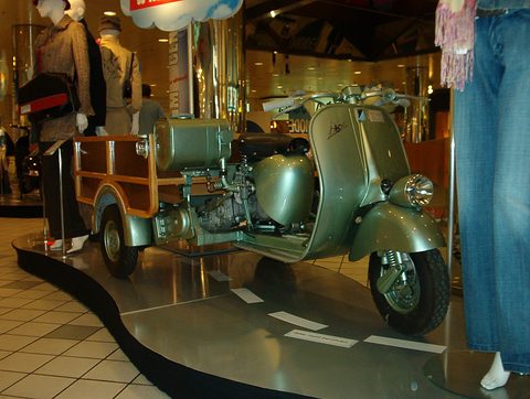 Grey-green scooter-fronted three-wheeler with a varnished wooden load bed, standing on a curved plinth among shop mannequins. Opens a larger view